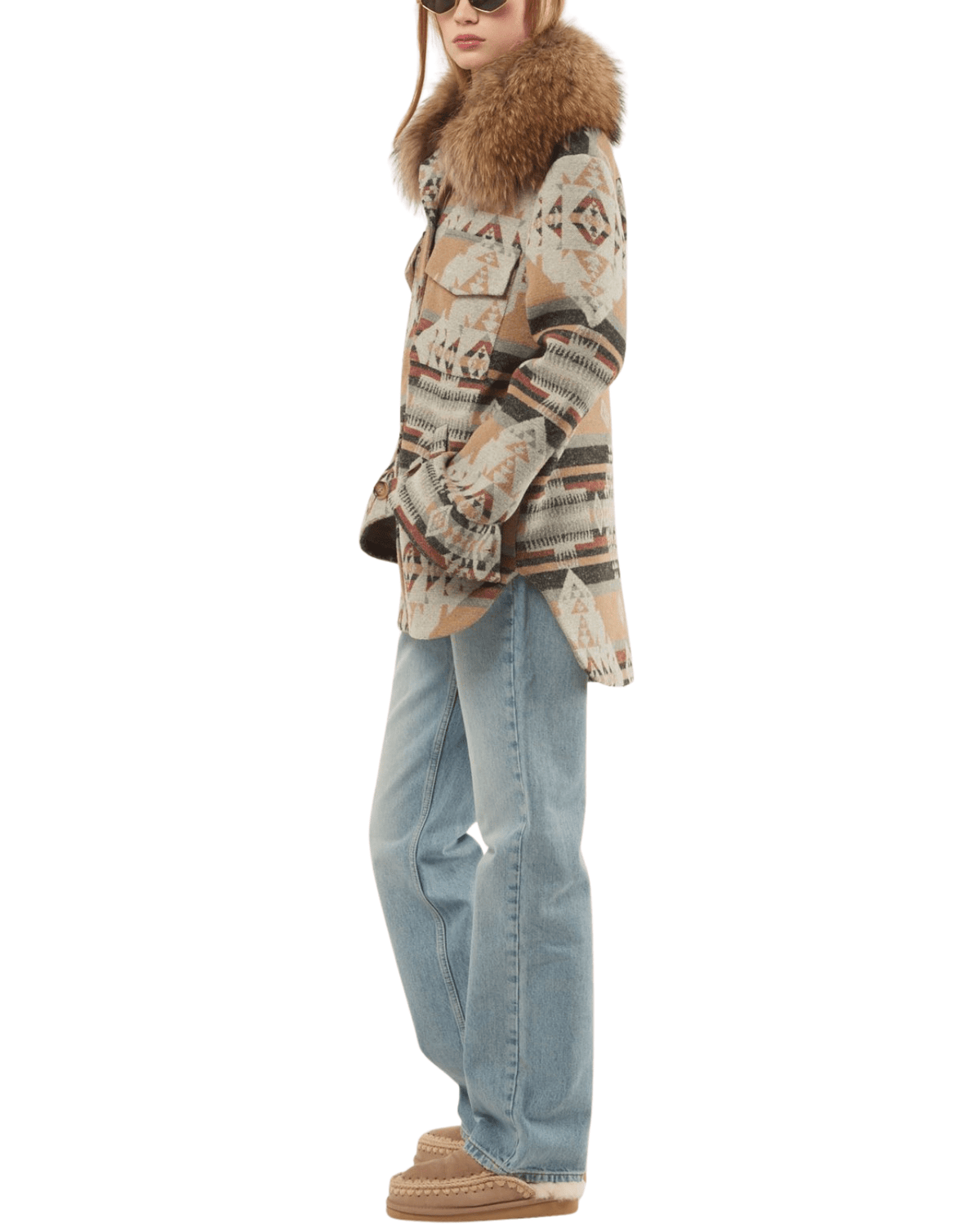 A woman modeling a patterned winter jacket with a large brown fur-trimmed collar, paired with light-wash blue jeans and brown mule shoes, shown against a plain white background.
