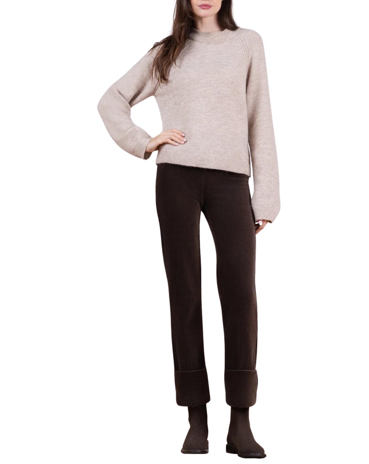 A woman modeling a light beige knit mock-neck sweater and dark brown cropped trousers with rolled cuffs, paired with dark brown ankle boots, against a plain white background.