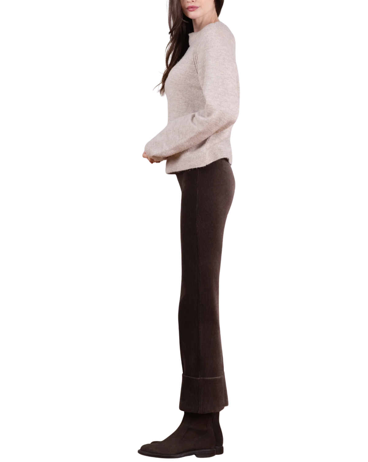 A woman shown in side profile wearing a light beige knit mock-neck sweater, dark brown flared pants, and dark brown ankle boots against a plain white background.