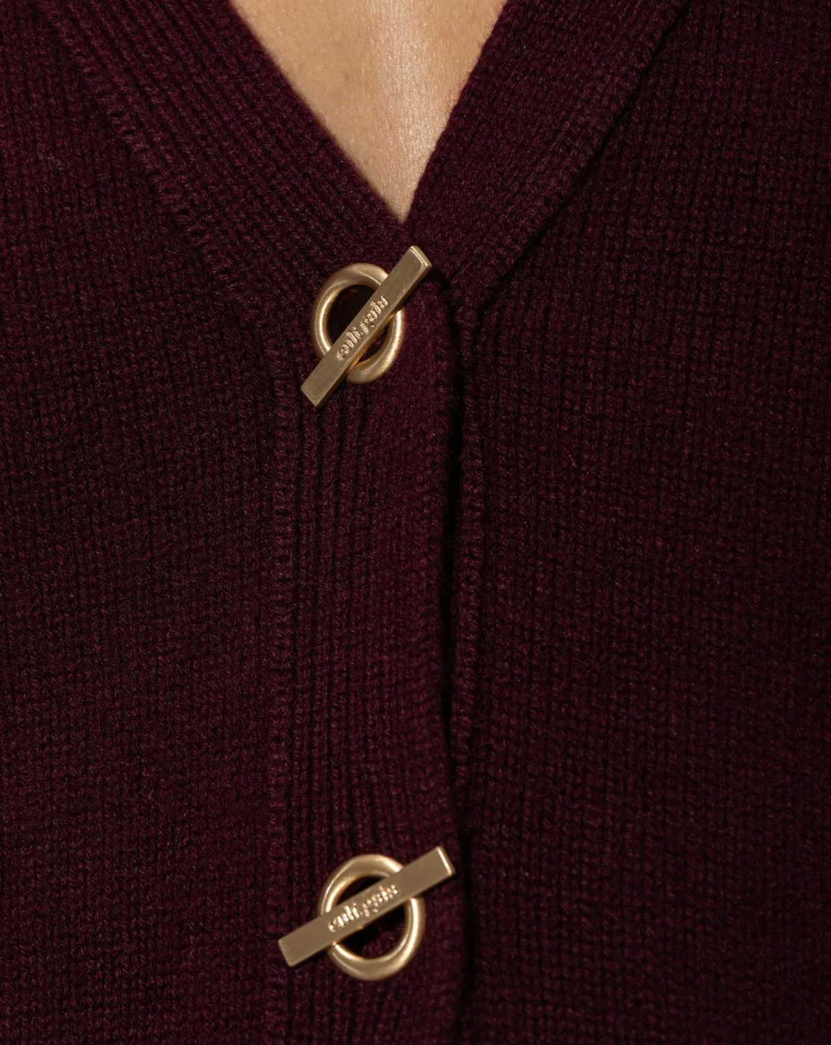 Close-up of a burgundy ribbed knit cardigan with gold-tone metal buttons featuring circular rings and engraved text.