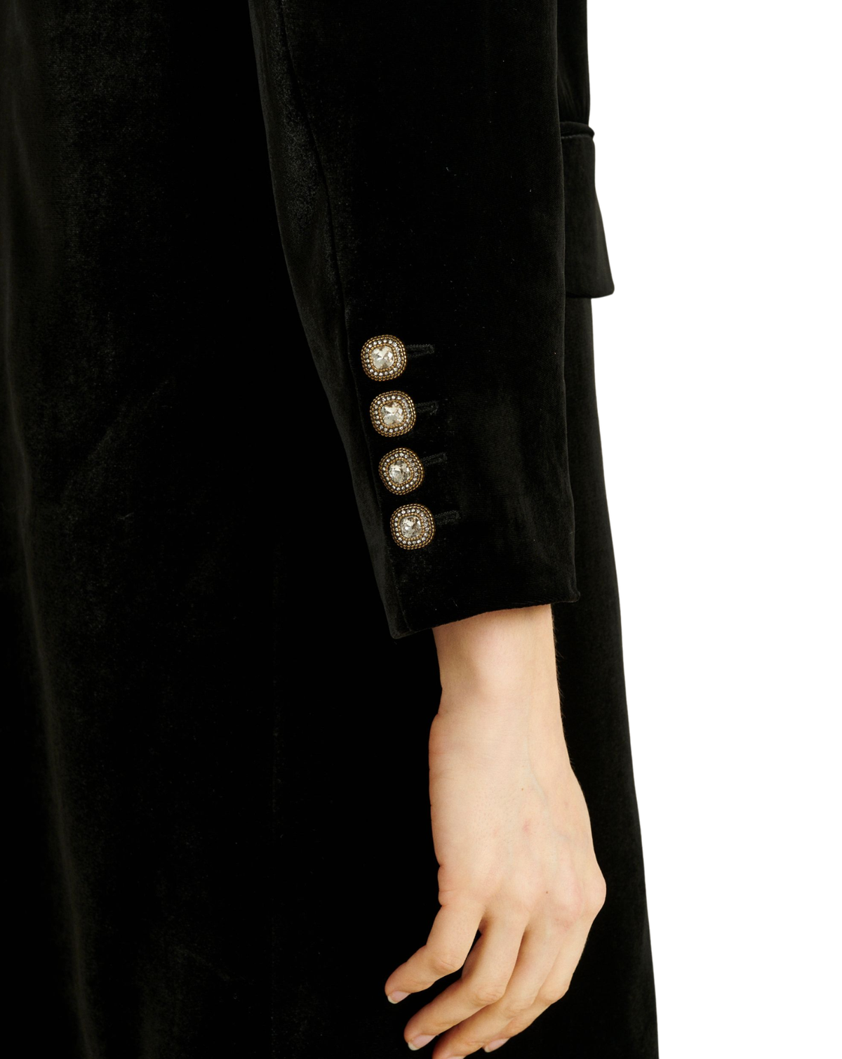 Close-up of a black velvet long coat sleeve with four decorative gold-toned buttons featuring white centers, worn by a model whose hand is visible against a plain white background.