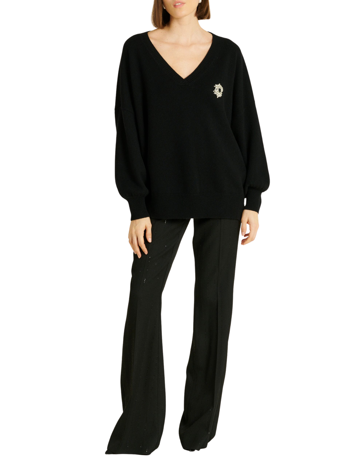 A model wearing a black knit sweater with a small white embroidered floral detail on the left chest, paired with black wide-leg trousers, against a plain white background.