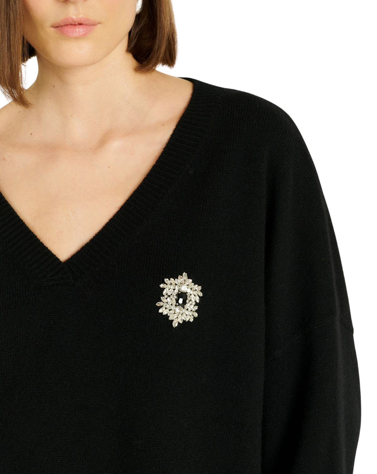 A close-up of a model wearing a black V-neck knit sweater with a decorative silver-toned brooch featuring clear rhinestones on the left chest, against a plain white background.