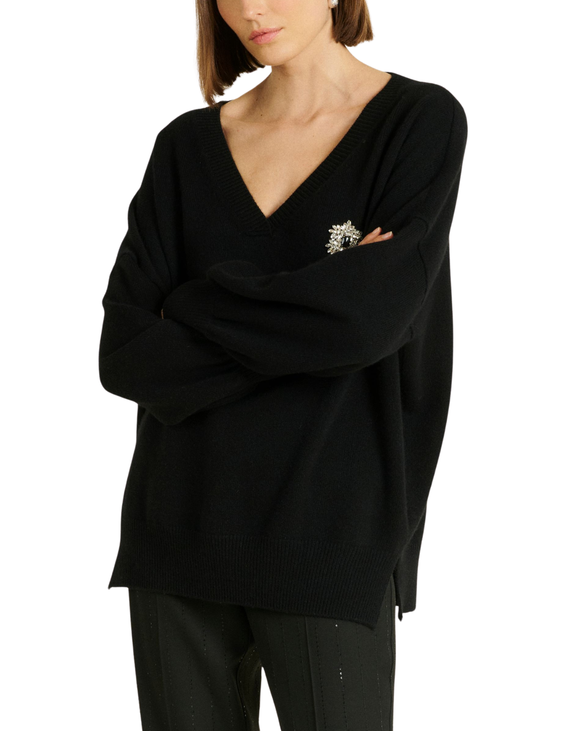 A woman wearing a black knit V-neck sweater with a decorative silver-toned brooch on the chest, paired with black pants, against a plain white background.