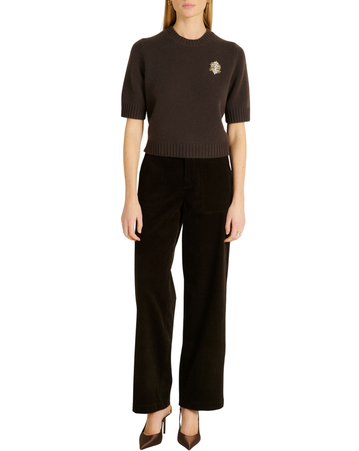 A model wearing a dark brown short-sleeve knit sweater with a small embroidered crest on the left chest, paired with dark brown wide-leg trousers and brown pointed-toe heels, against a plain white background.