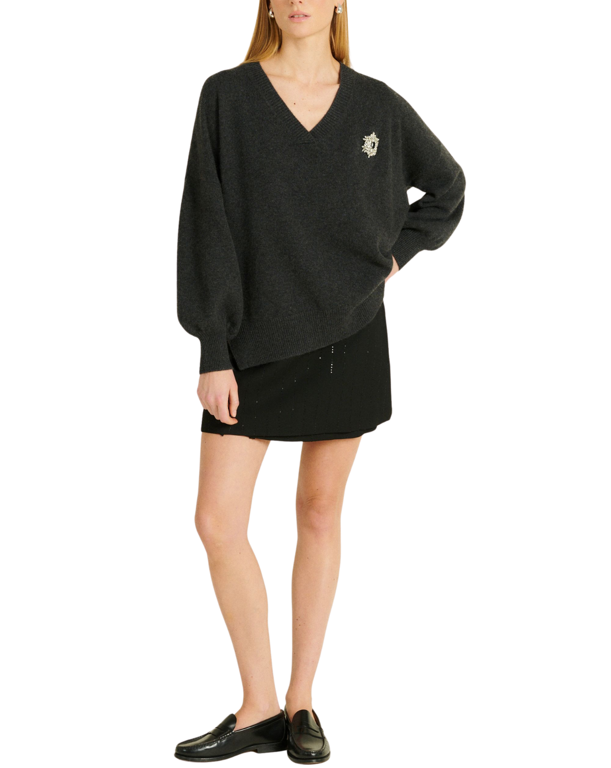 A model wearing a dark charcoal gray knit sweater dress with a small embroidered crest on the left chest, paired with a black mini skirt and black loafers, against a plain white background.