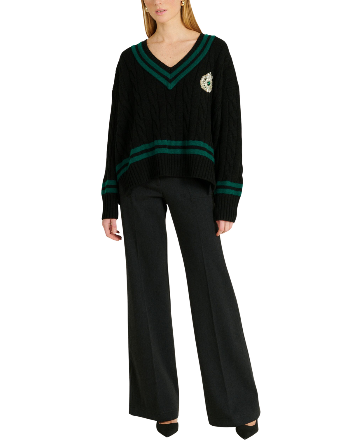 A woman modeling a black cable-knit sweater with green stripes and a crest emblem on the chest, paired with black wide-leg trousers, against a plain white background.