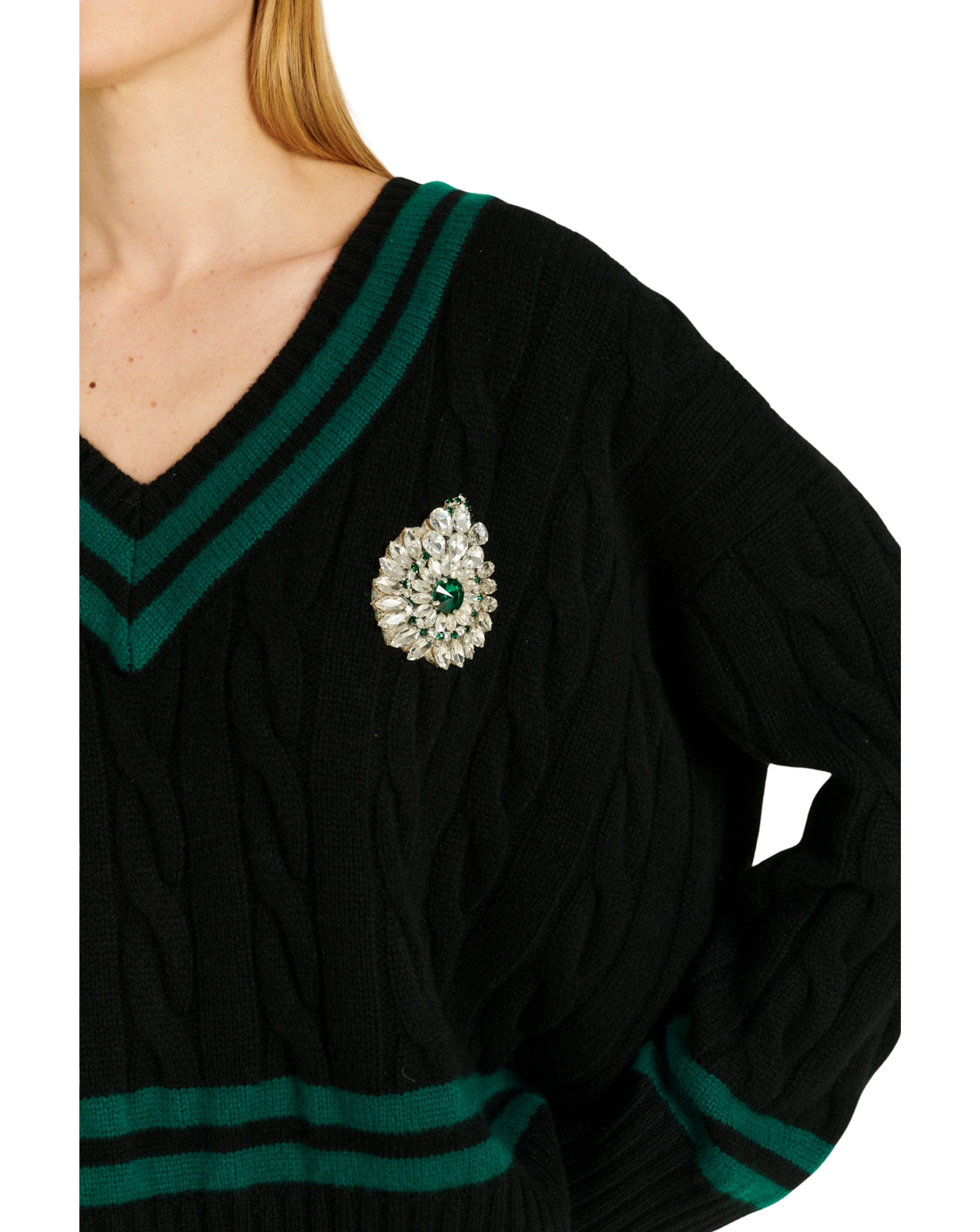 A close-up of a person wearing a black cable-knit V-neck sweater with dark green stripes on the collar and hem, featuring a jeweled brooch on the chest.