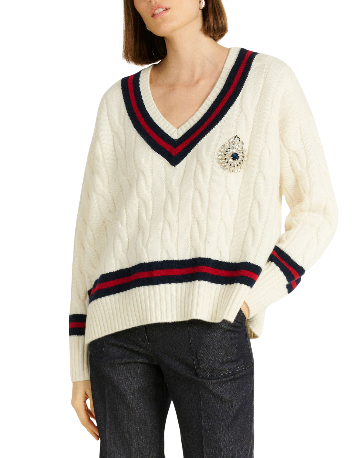 A woman wearing a cream cable-knit V-neck sweater with navy and red stripes on the collar and cuffs, featuring a jeweled brooch on the chest, paired with dark gray pants against a white background.