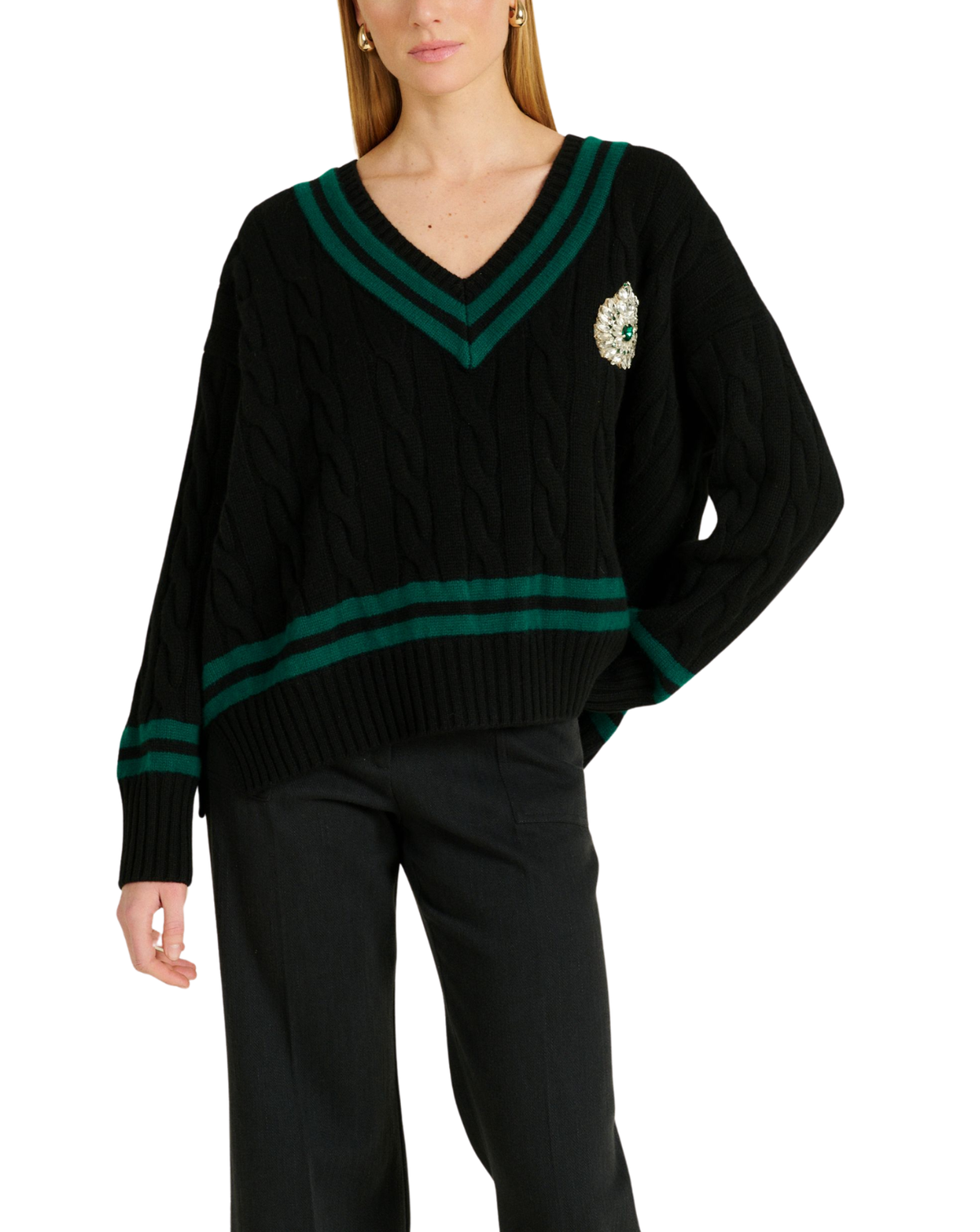 A woman wearing a black cable-knit V-neck sweater with dark green stripe accents on the collar and cuffs, and a decorative crest patch on the left chest, paired with black pants against a plain white background.