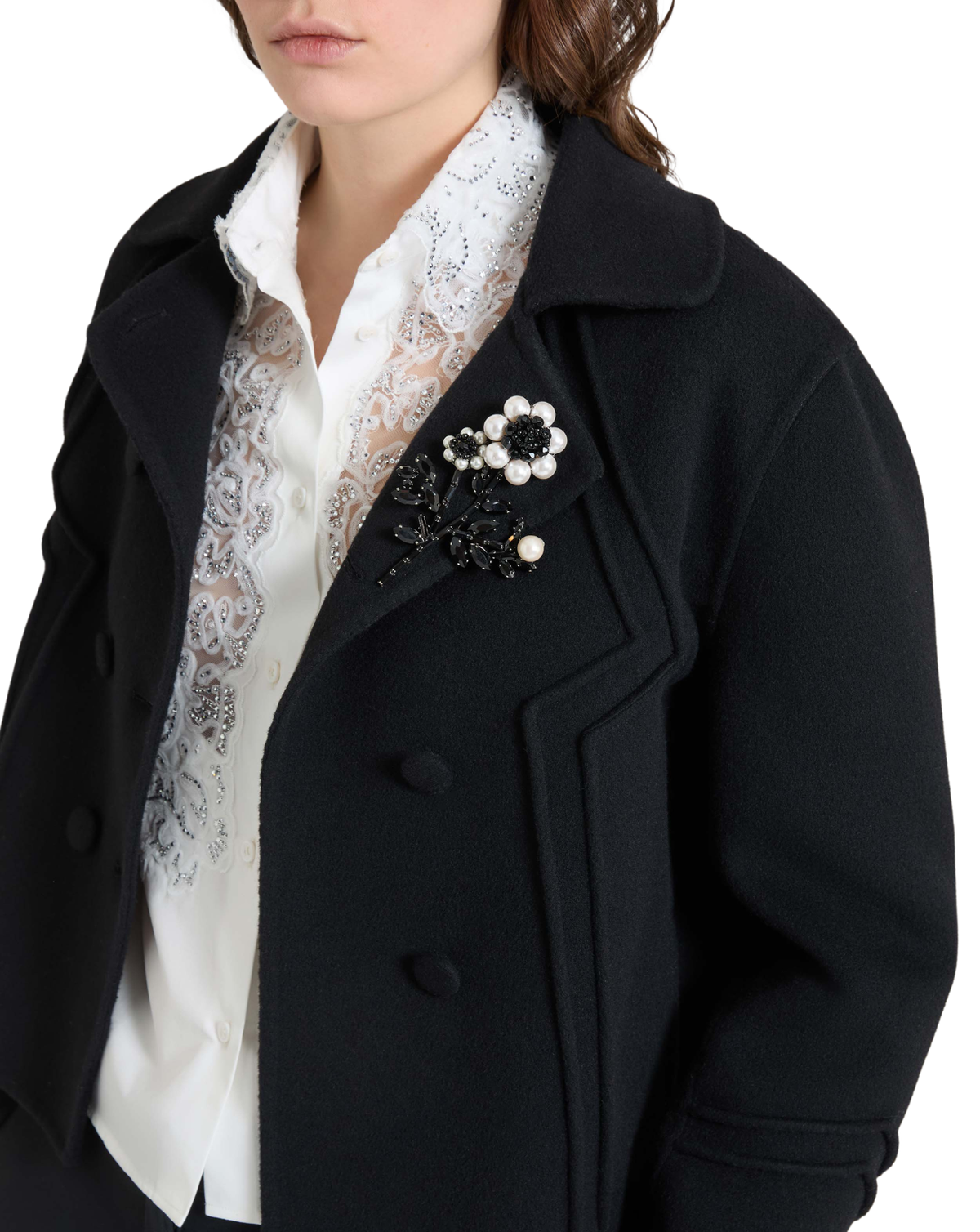 A close-up of a model wearing a black double-breasted coat over a white lace blouse, featuring a decorative black and white floral brooch on the lapel.