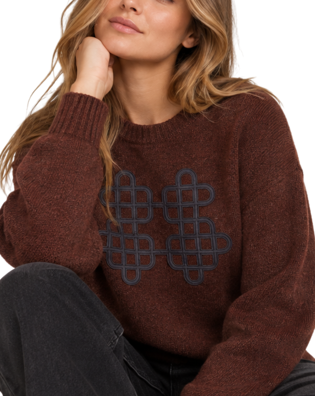 A woman wearing a dark brown knit sweater with a large dark navy geometric pattern on the chest, paired with dark gray jeans, against a plain white background.