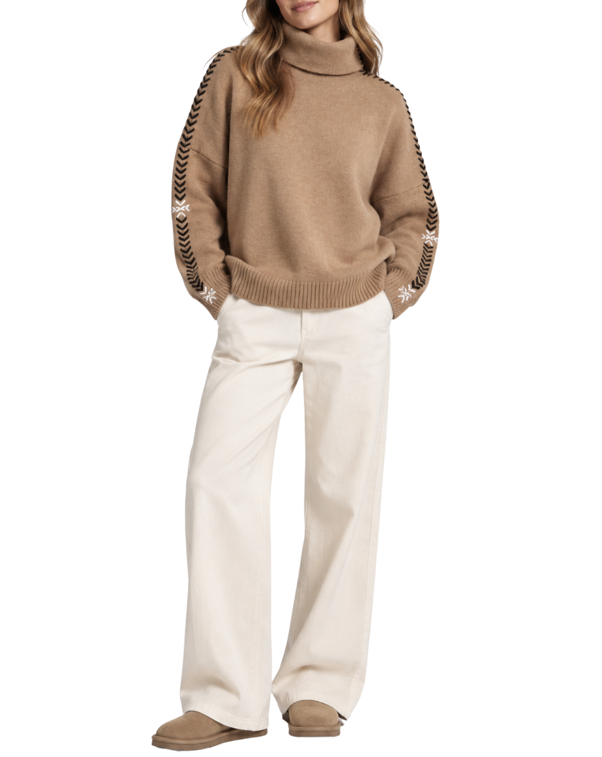 A model wearing a camel brown turtleneck sweater with black and white patterned knit sleeve stripes, paired with off-white wide-leg trousers and tan slippers, set against a plain white background.