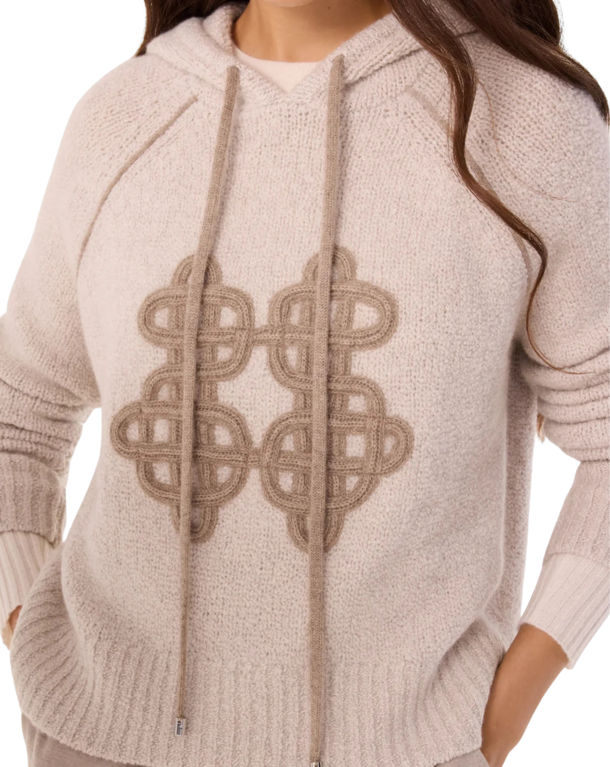 A close-up of a woman wearing a chunky knit hoodie in a light oatmeal beige color, featuring a large geometric interlocking pattern on the front and matching drawstrings.