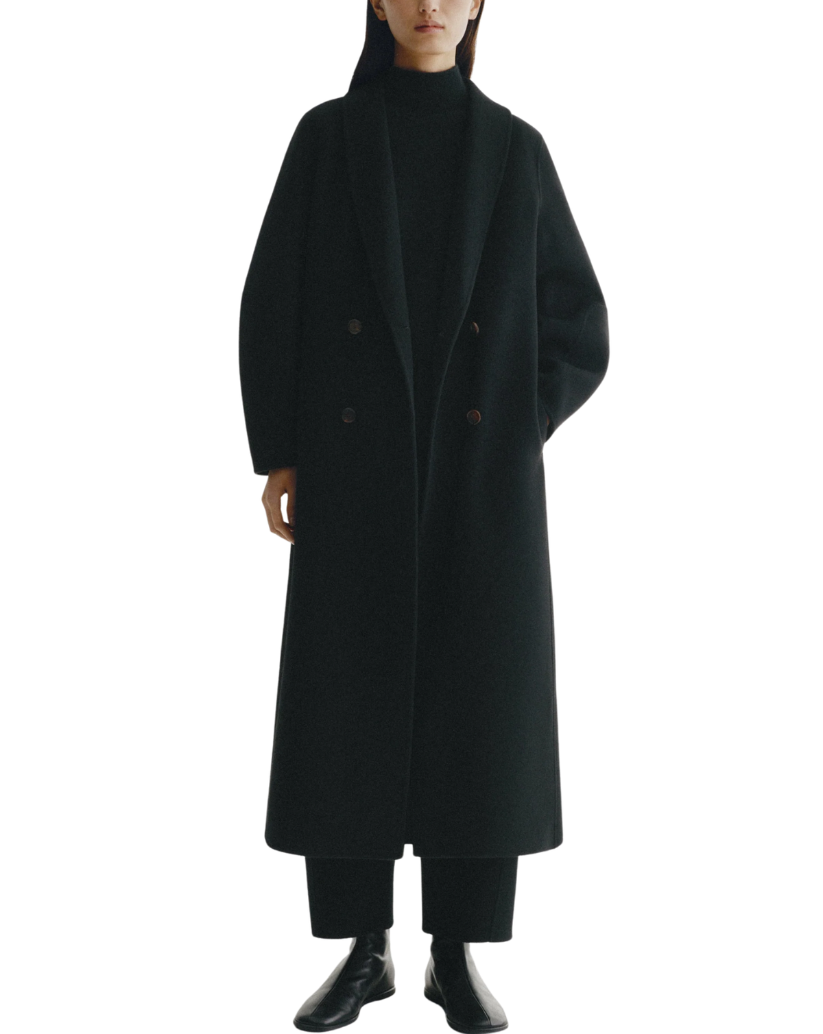 A model wearing a long, double-breasted black coat over a black turtleneck, black trousers, and black leather boots, shot against a solid white background.