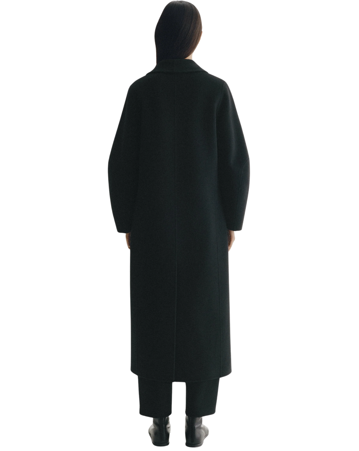 A model viewed from the back wearing a long black coat, black trousers, and black shoes against a plain white background.