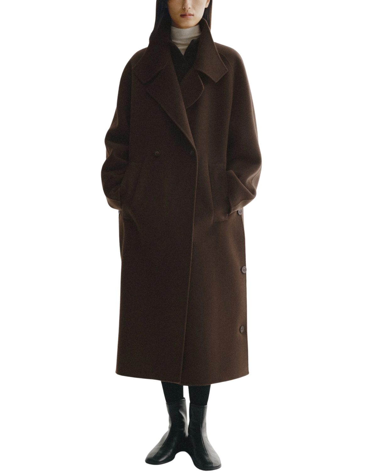 A model wearing a long, dark brown double-breasted coat over a black outfit and black boots, set against a plain white background.