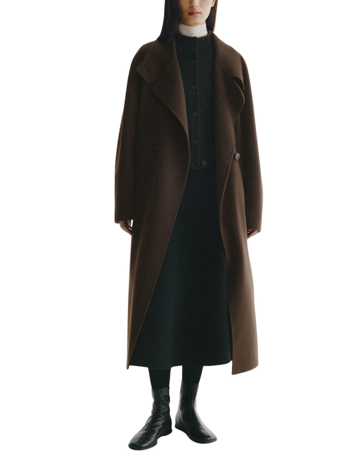 A model wearing a long brown wool-blend coat over a black dress, black top, black boots, and a white collar, photographed against a solid white background.