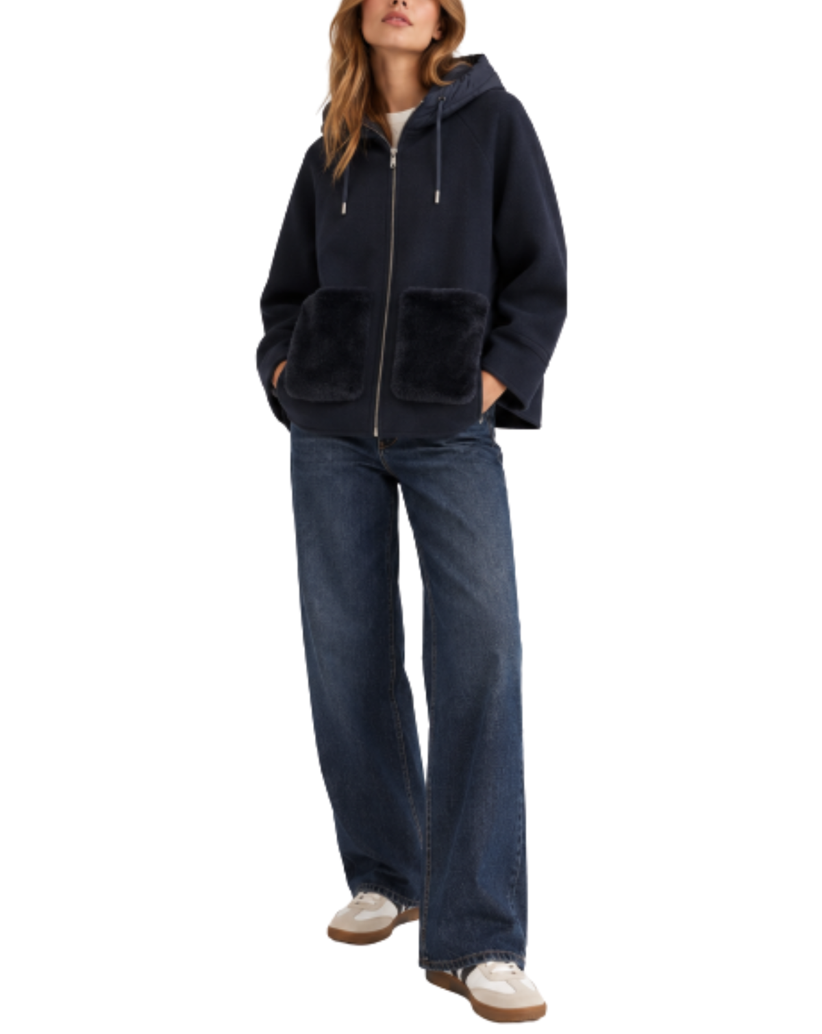 A woman modeling a navy blue zip-up hooded jacket with fuzzy faux-fur trim on the hood and front patch pockets, paired with dark blue wide-leg jeans and white sneakers, against a plain white background.