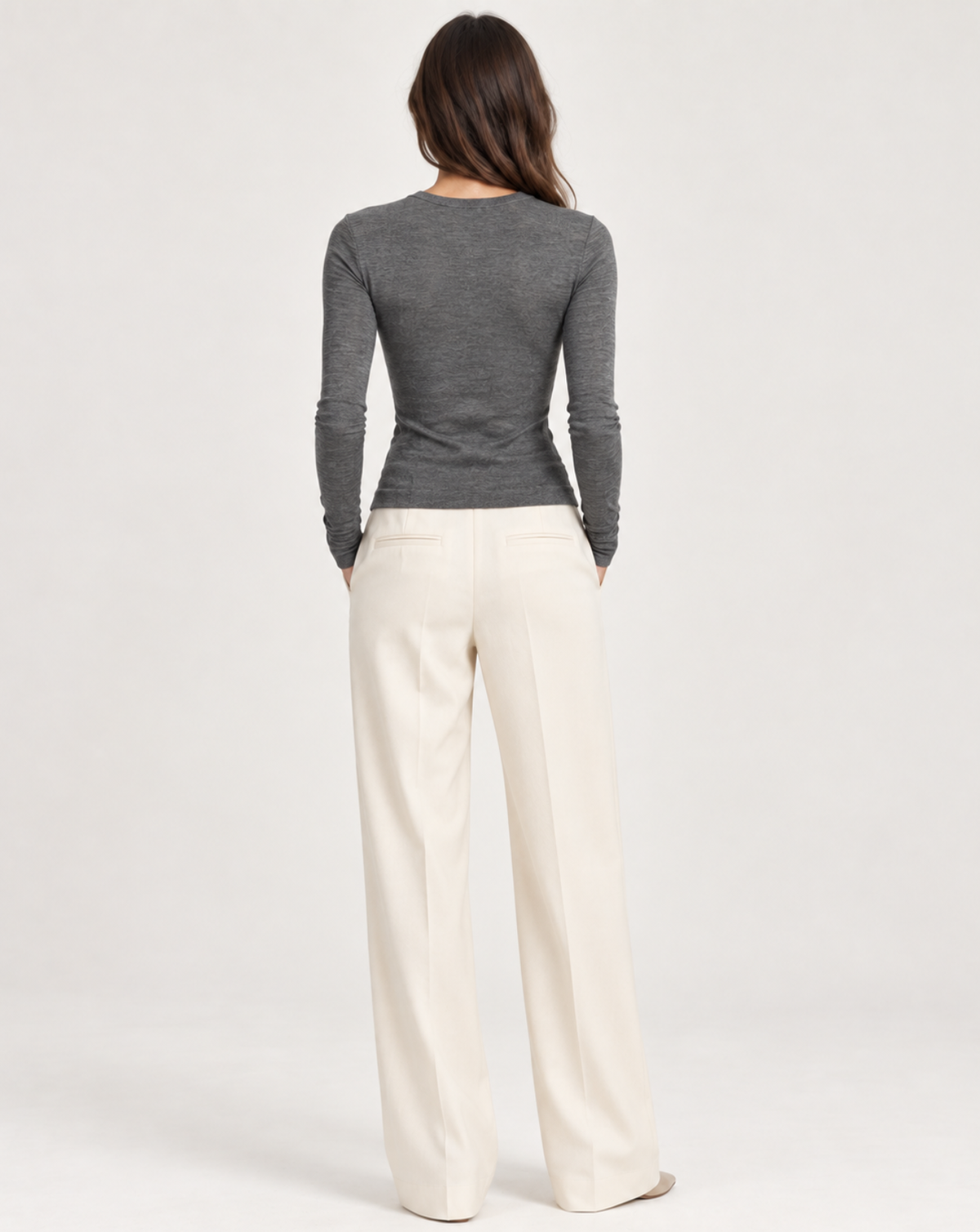 A woman shown from the back wearing a fitted dark gray long-sleeve top and off-white wide-leg trousers, photographed against a clean light gray studio background.