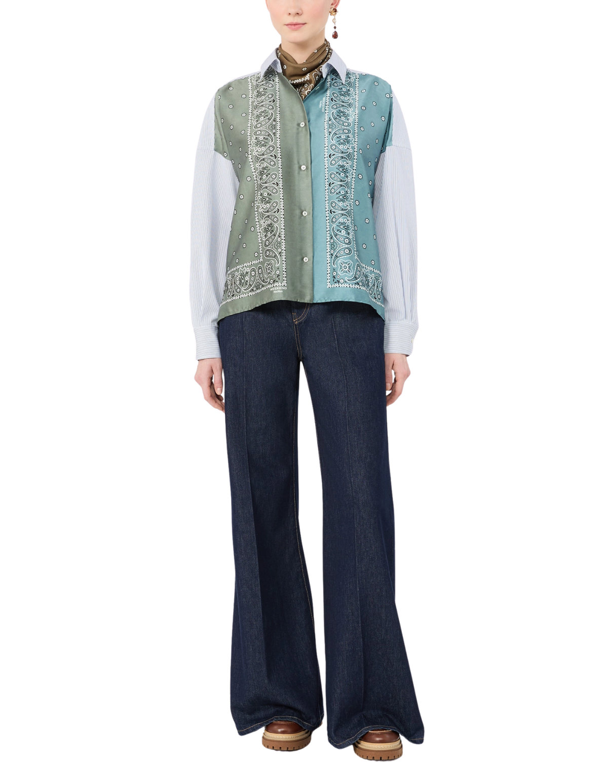 A model wearing a sleeveless button-front vest with a mixed print design, featuring sage green and light blue panels with white paisley patterns, styled with a white shirt, brown scarf, dark blue wide-leg jeans, and brown shoes against a plain white background.