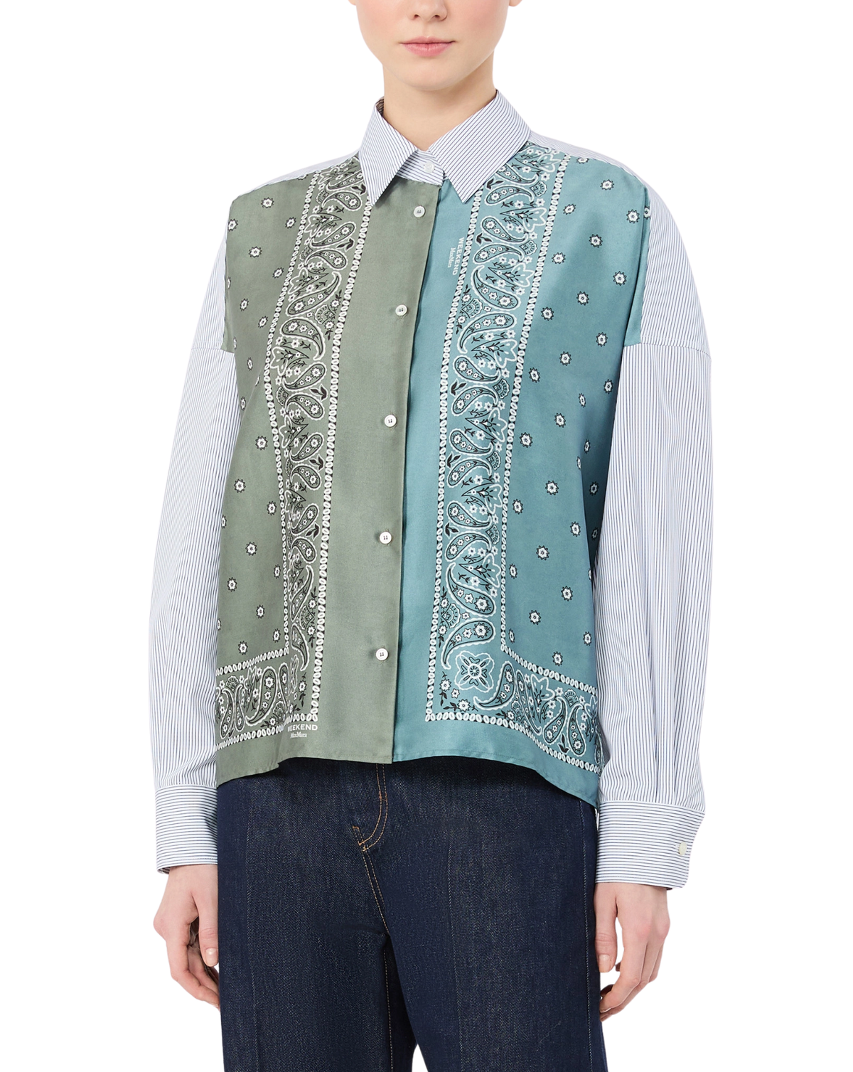 A model wearing a sleeveless button-front vest with a color-blocked design in sage green and light blue, featuring white paisley and dot patterns, layered over a light blue striped button-up shirt and dark blue jeans.