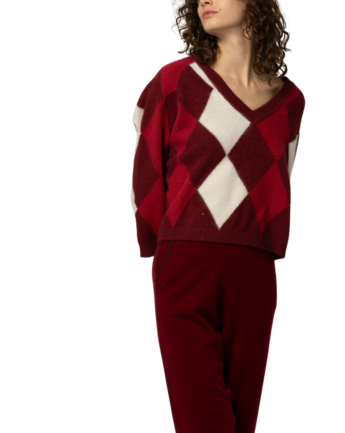 A model wearing a cropped knit sweater with a red and cream argyle pattern, paired with matching dark red sweatpants, shot against a solid white background.