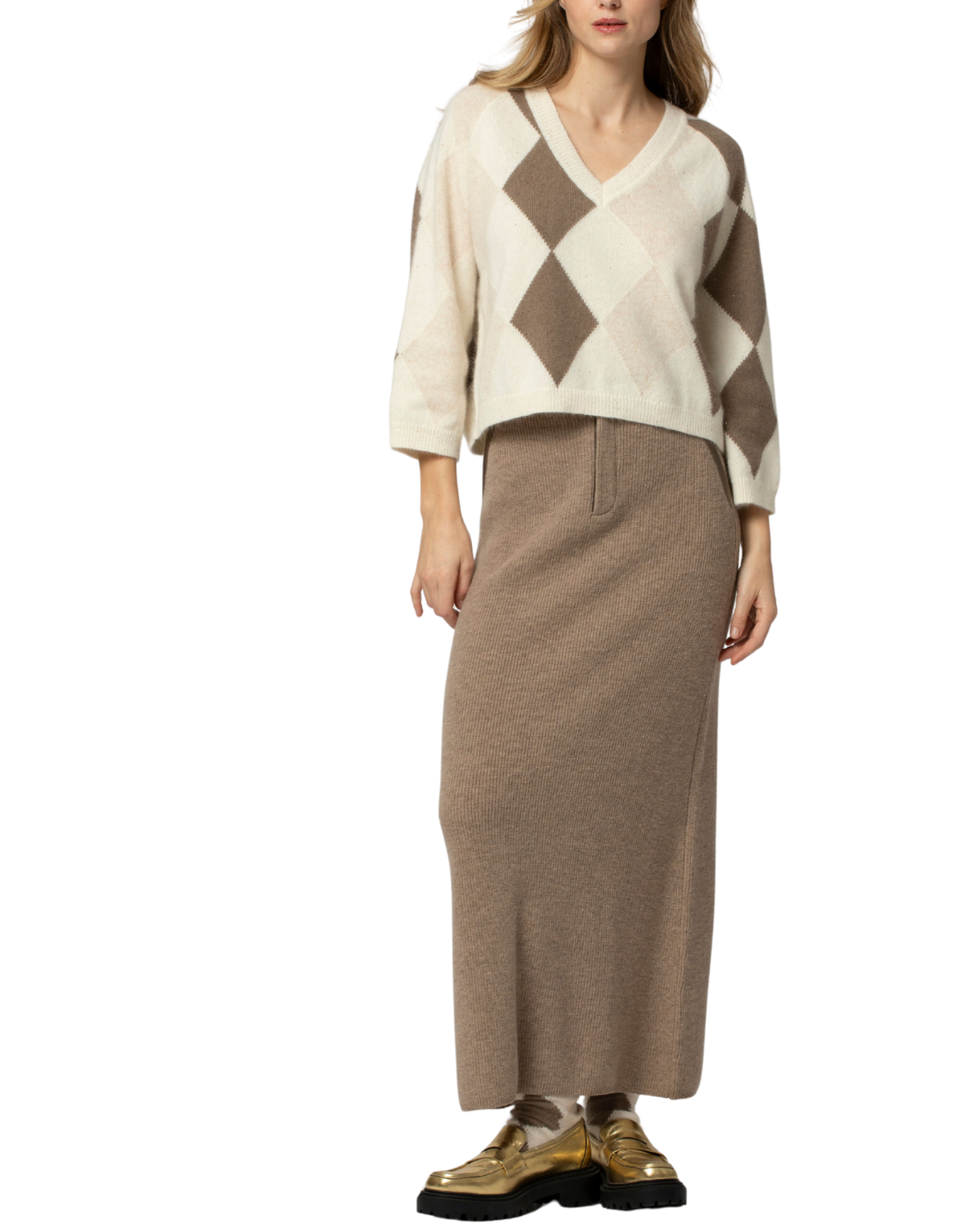 A model wearing a cream and brown argyle knit sweater vest over a taupe knit midi skirt, paired with metallic gold loafers, shot against a plain white background.