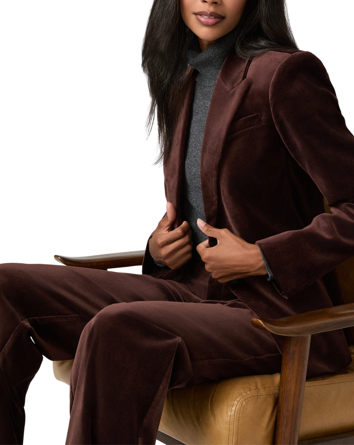 A woman seated in a tan leather chair wearing a matching dark brown velvet blazer and trousers set over a gray turtleneck sweater, photographed against a clean white studio background.