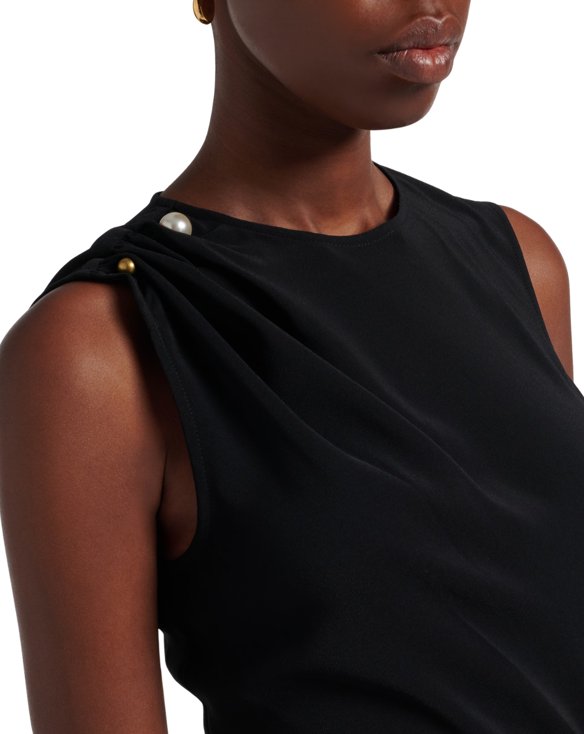 Close-up of a model wearing a black asymmetrical draped sleeveless top with gold and pearl button details on the shoulder, set against a plain white background.