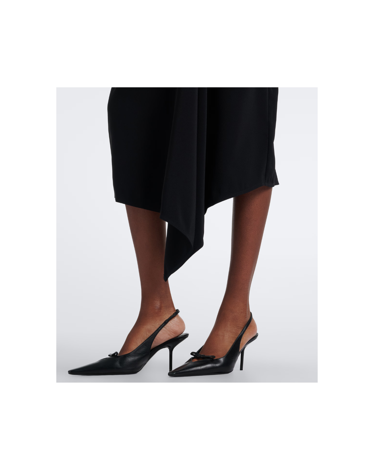 Black pointed-toe slingback heels with a small bow detail on the strap, shown on a model wearing a black asymmetrical skirt against a light gray background.