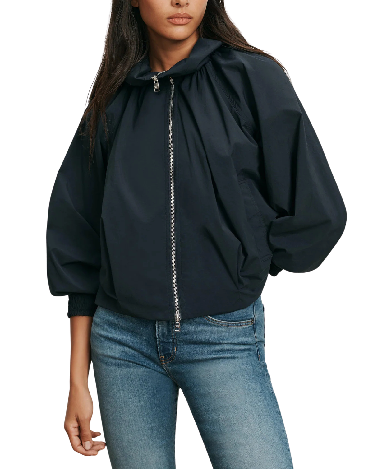 A woman wearing a dark navy hooded bomber jacket with a silver zipper and voluminous sleeves, paired with blue jeans, against a plain white background.