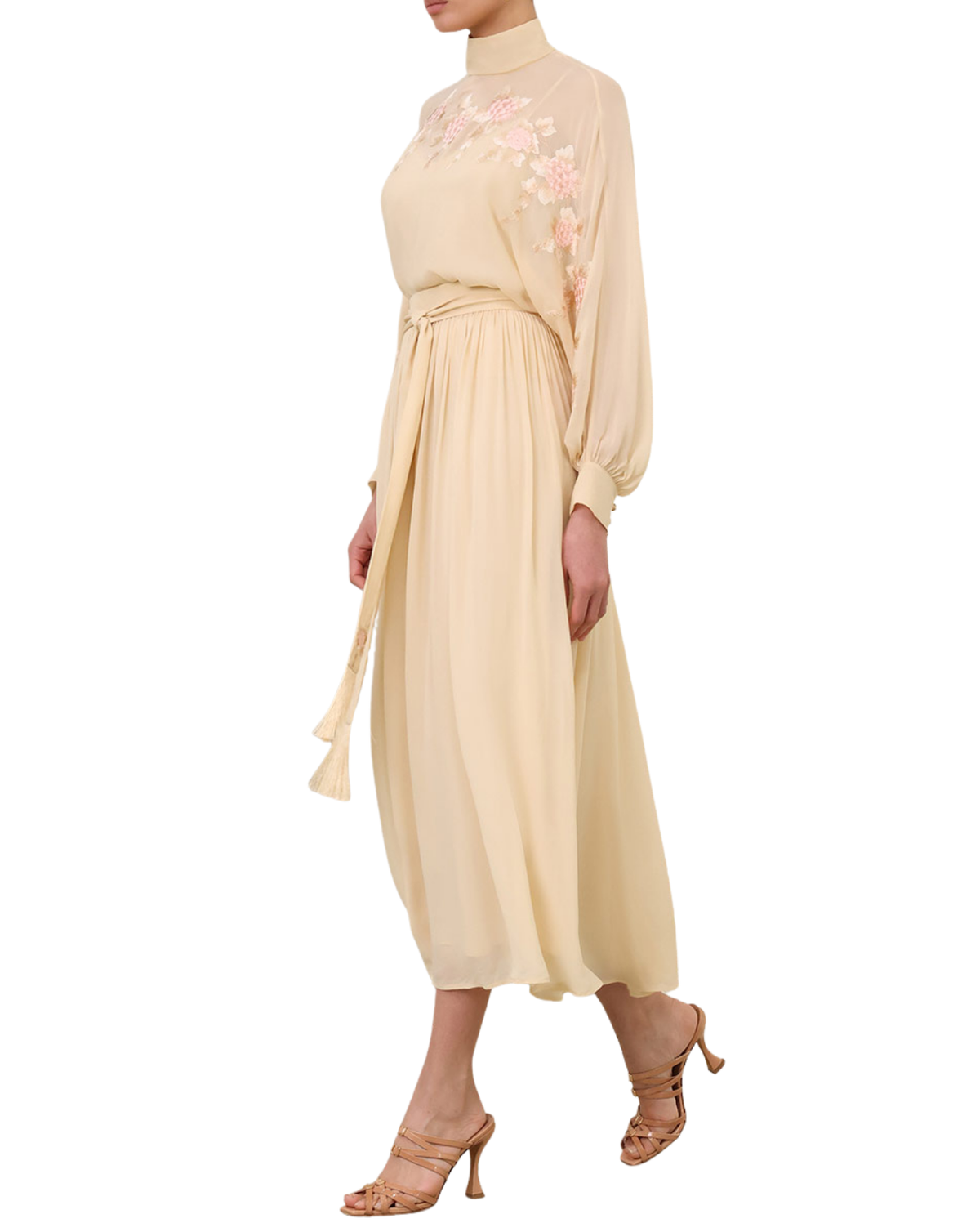 A model wearing a high-neck, long-sleeved midi dress in a light cream or pale beige color, decorated with pink floral embroidery on the upper chest and shoulder area. The dress features a tie-waist belt and a flowing skirt, paired with beige strappy high-heeled sandals against a plain white background.