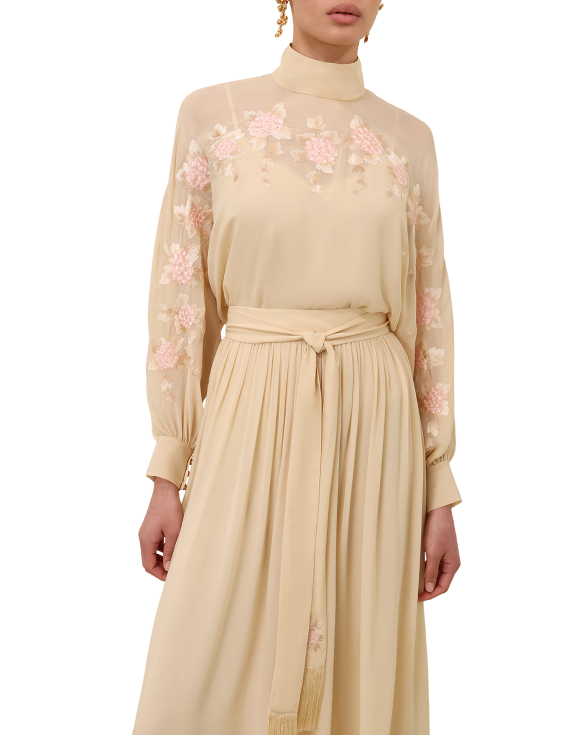 A model wearing a beige high-neck long-sleeve dress with pink floral embroidery on the chest and sleeves, featuring a matching fabric tie belt at the waist.