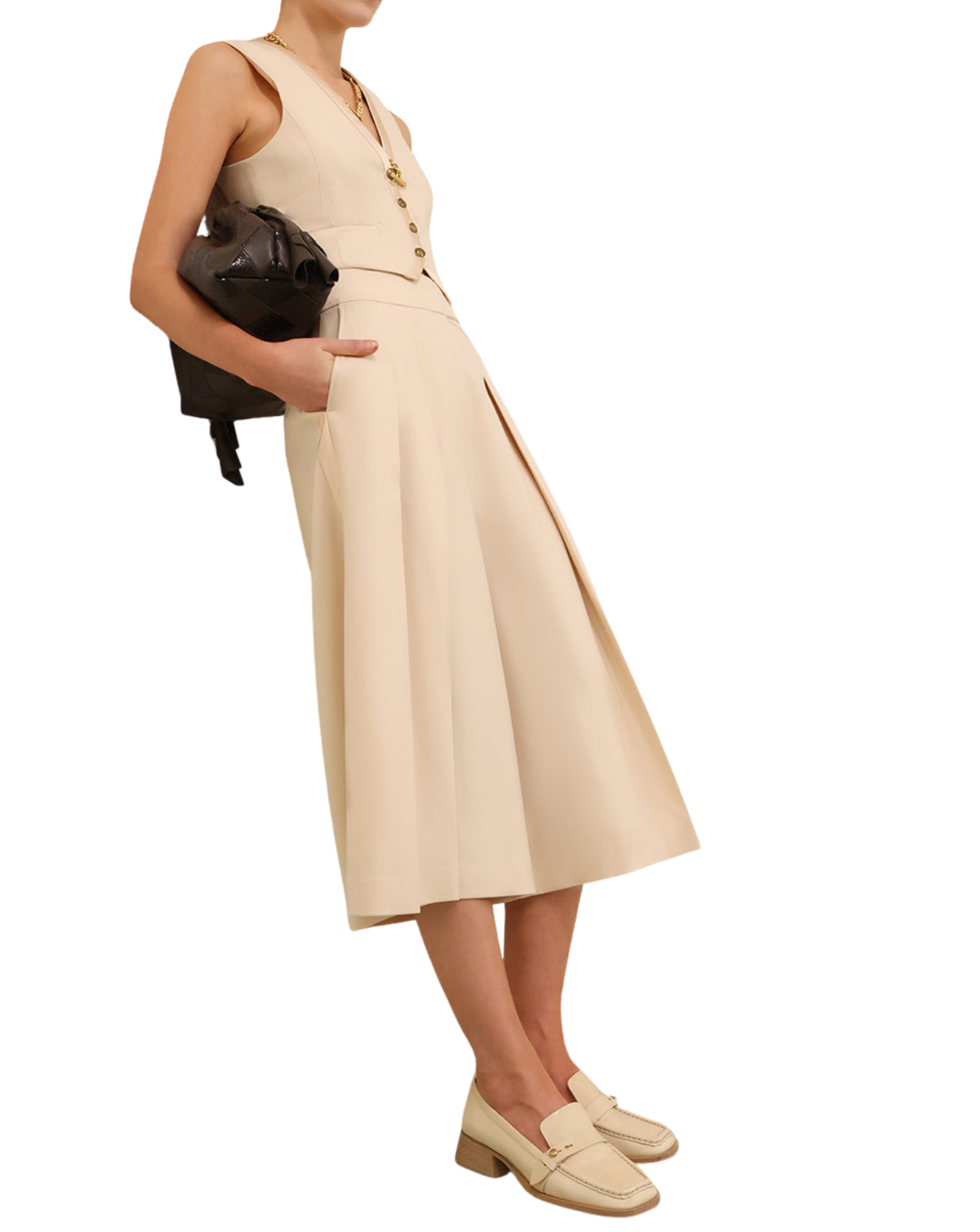 A model wearing a sleeveless beige midi dress with a buttoned waist, holding a black handbag and wearing beige loafers, set against a plain white background.