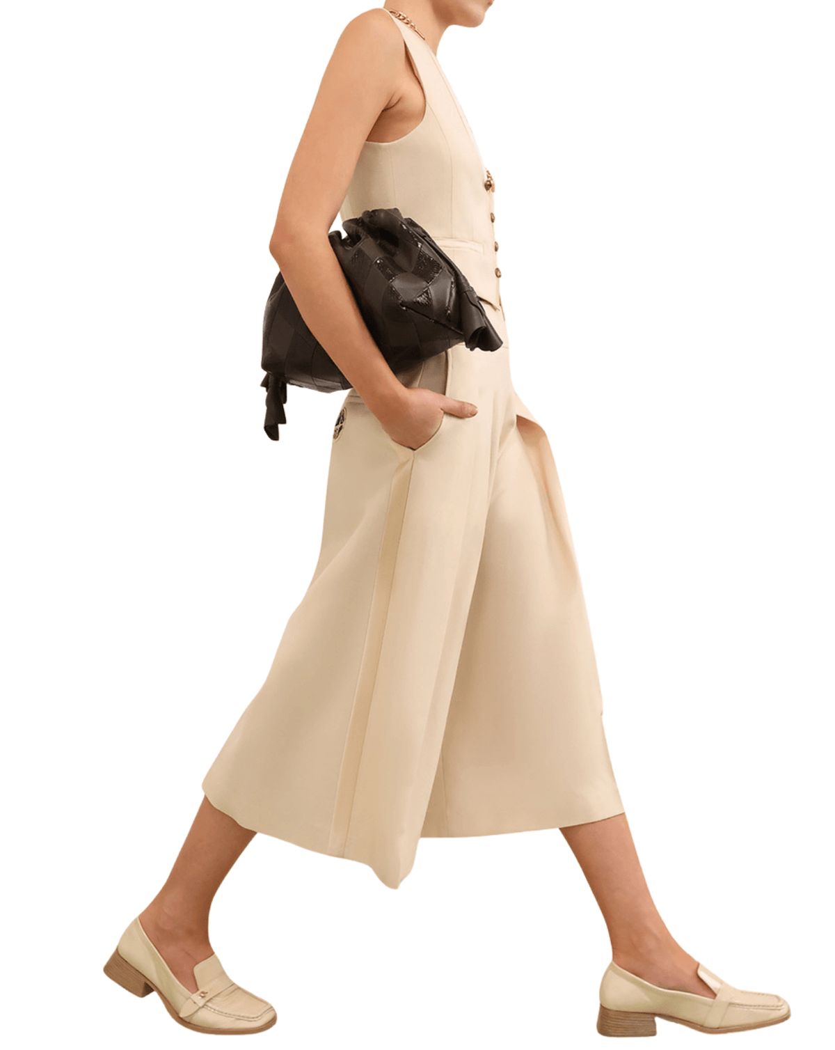 A model is shown from the side wearing a sleeveless, high-waisted cream-colored jumpsuit with wide legs, paired with a black leather shoulder bag and matching cream loafers, set against a plain white background.