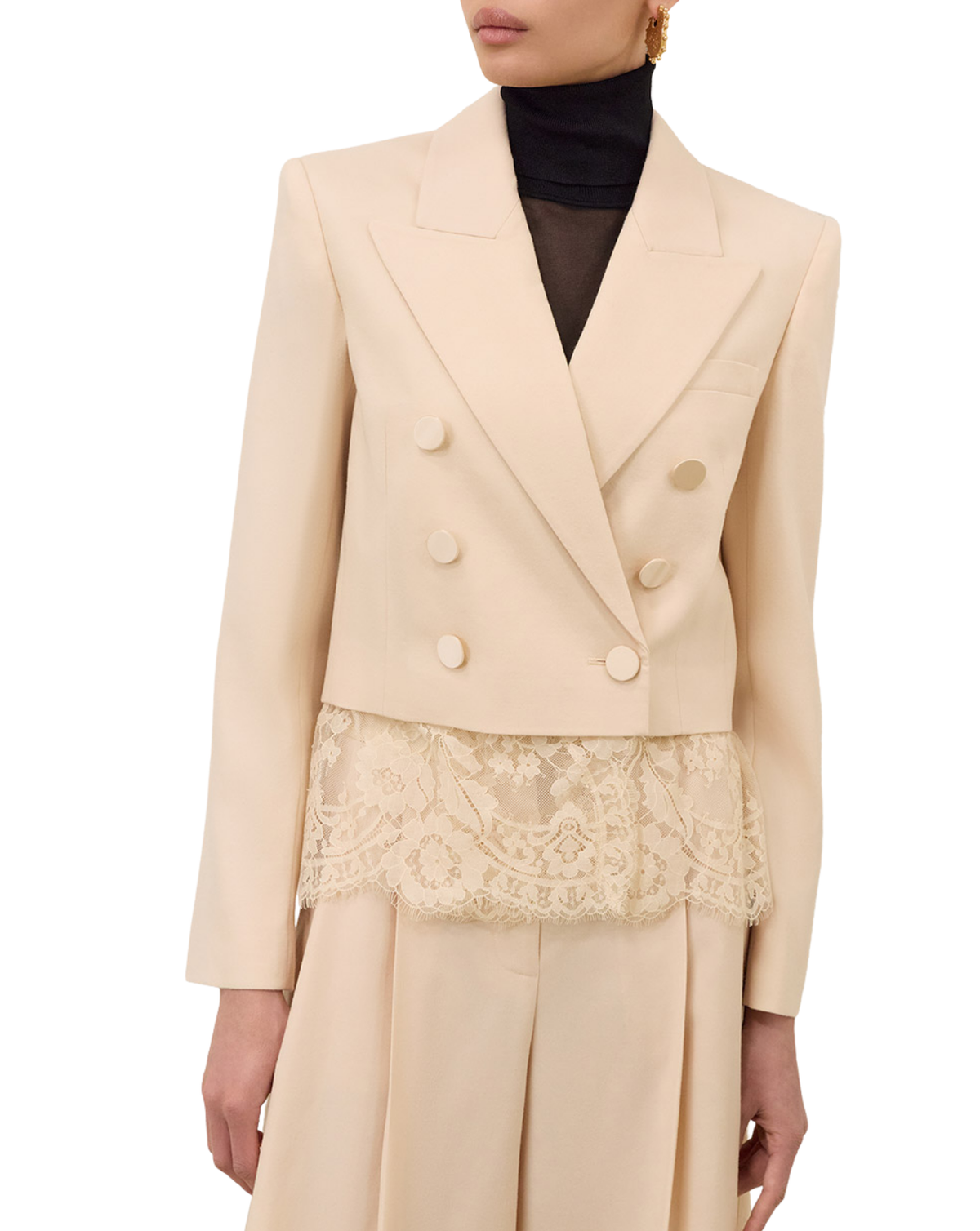 A model wearing a structured cream-colored double-breasted blazer with a matching pleated skirt featuring a lace overlay, styled with a black turtleneck.
