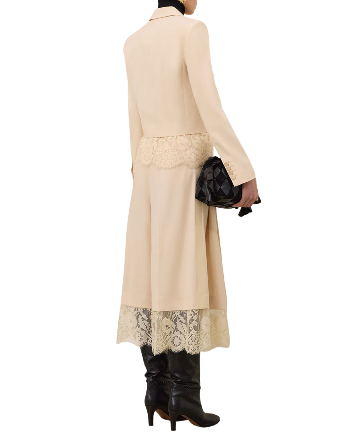A model shown from the back wearing a long beige coat with lace trim at the waist and hem, paired with black leather boots and holding a black patterned clutch bag against a plain white background.