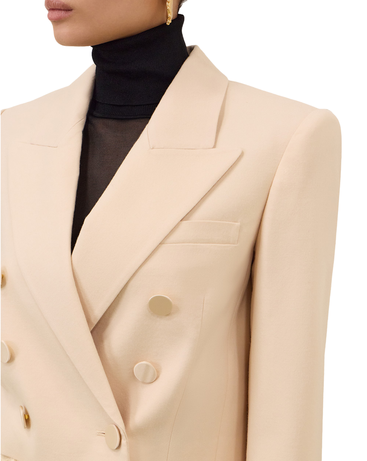 A close-up of a model wearing a structured cream-colored double-breasted blazer over a black turtleneck, with a gold earring visible.