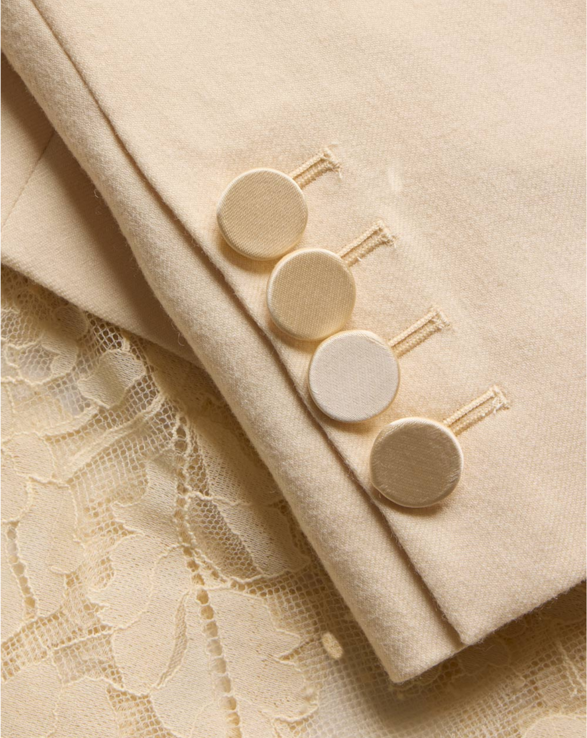 Close-up of a cream-colored wool blazer sleeve with four round buttons and a matching cream lace fabric underneath.