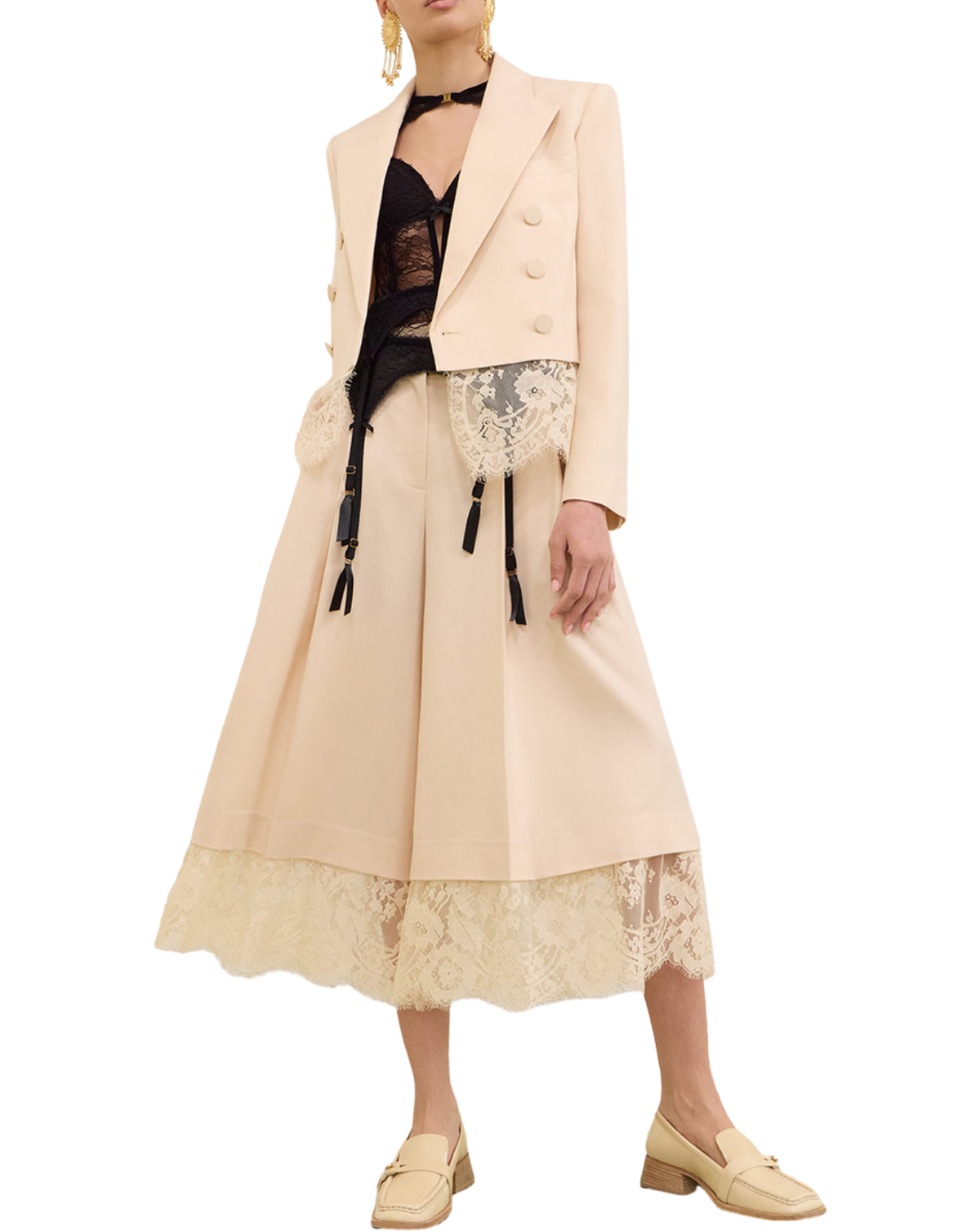 A model wears a beige double-breasted blazer with black tassels, a matching beige midi skirt with a lace hem, a black lace top, and beige loafers, set against a plain white background.
