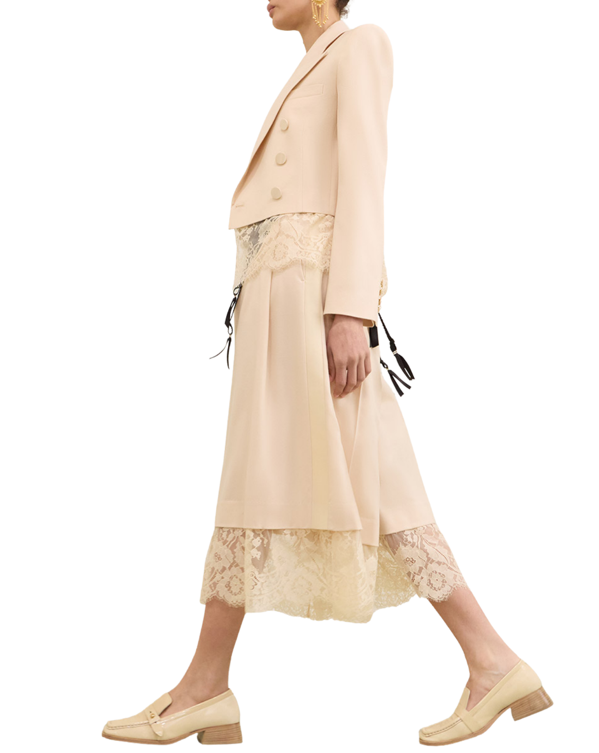 A model shown from the side wearing a light beige double-breasted blazer, a matching midi skirt with lace trim, and beige loafers against a plain white background.