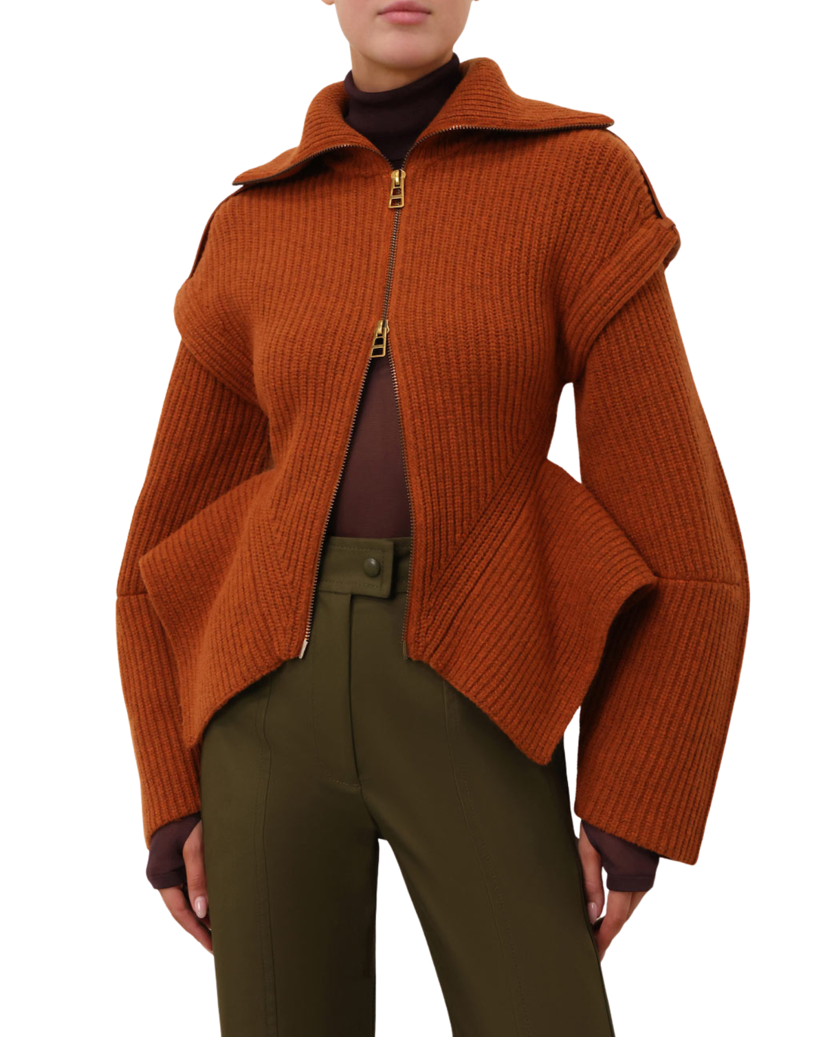 A model wearing a rust-orange ribbed knit asymmetrical zip jacket with a high collar, styled with olive green trousers and a dark brown turtleneck against a white background.