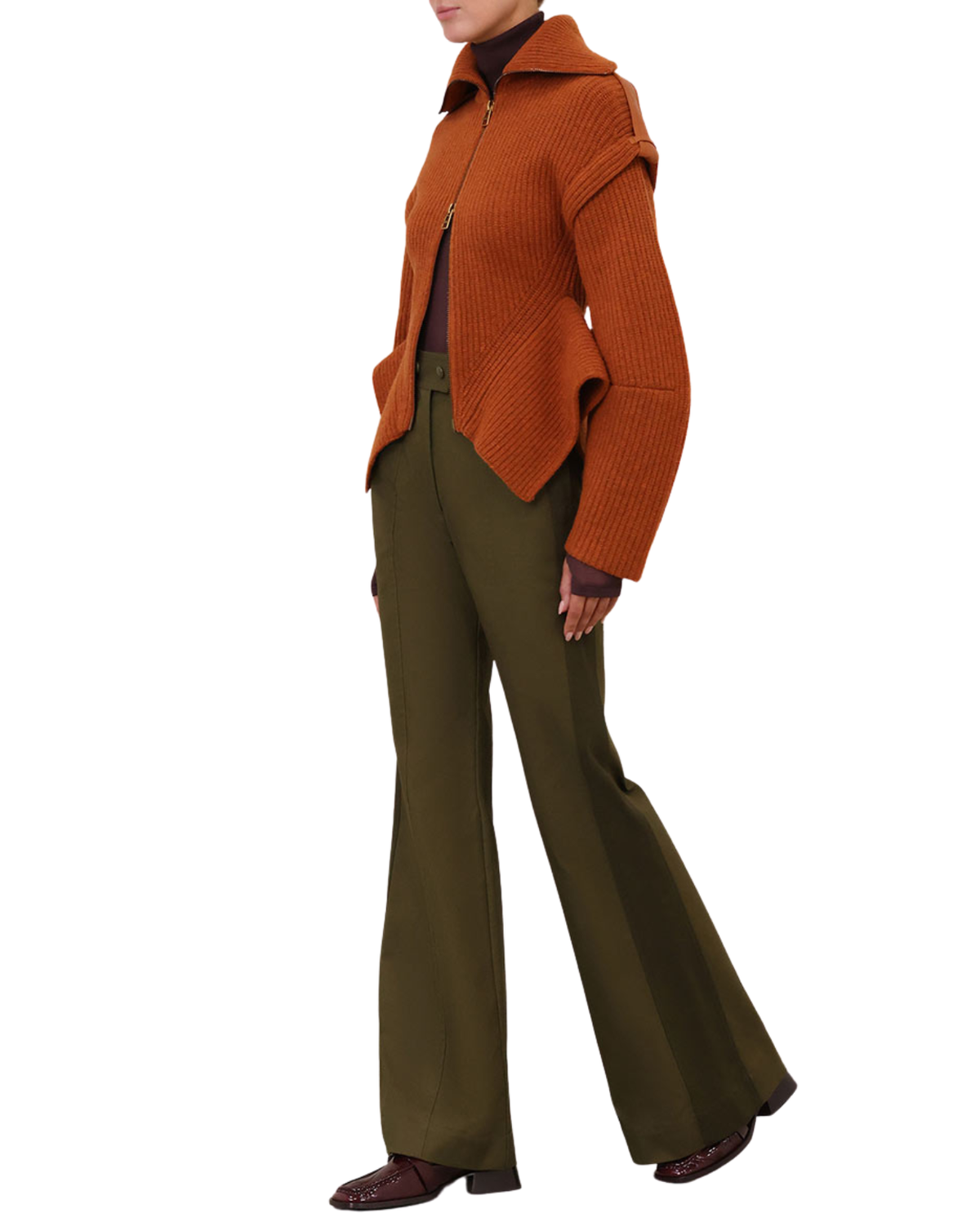 A model is shown from the neck down wearing a rust-orange ribbed knit zip-up cardigan, olive green flared trousers, and dark brown shoes, set against a plain white background.