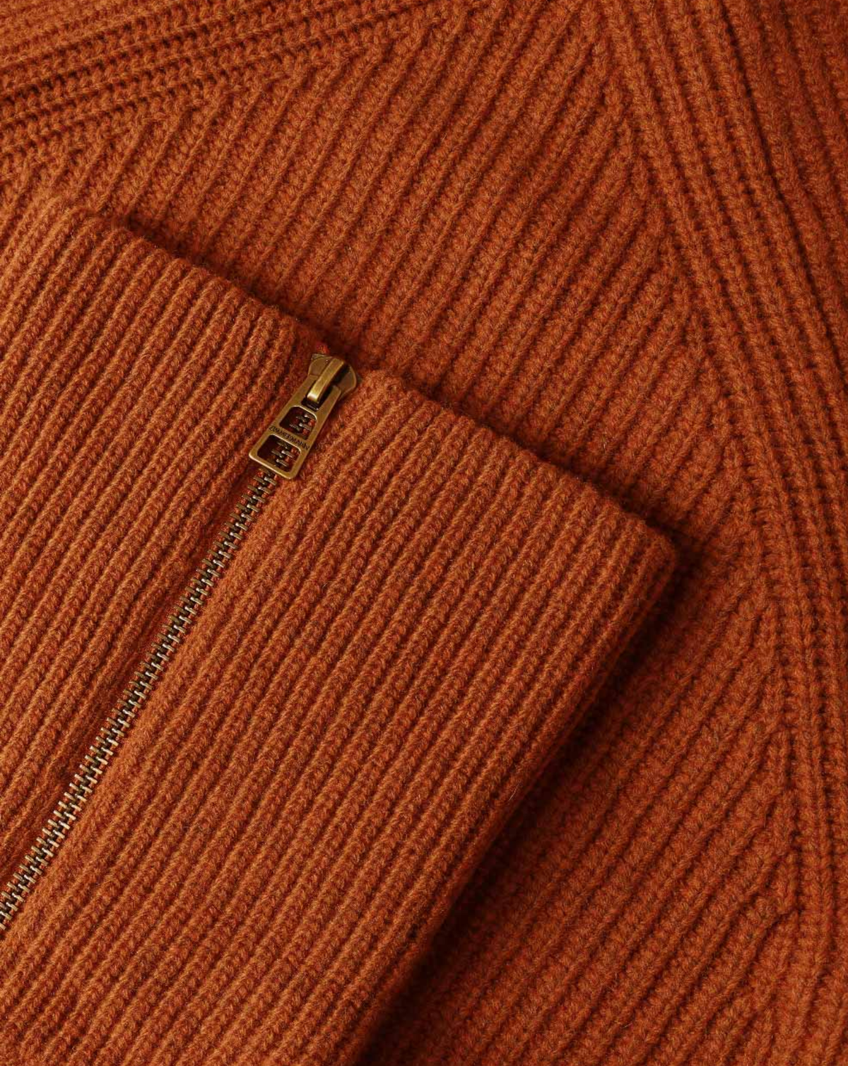 Close-up of a ribbed knit sweater in a warm rust orange color, showing a folded cuff with a gold-tone metal zipper.