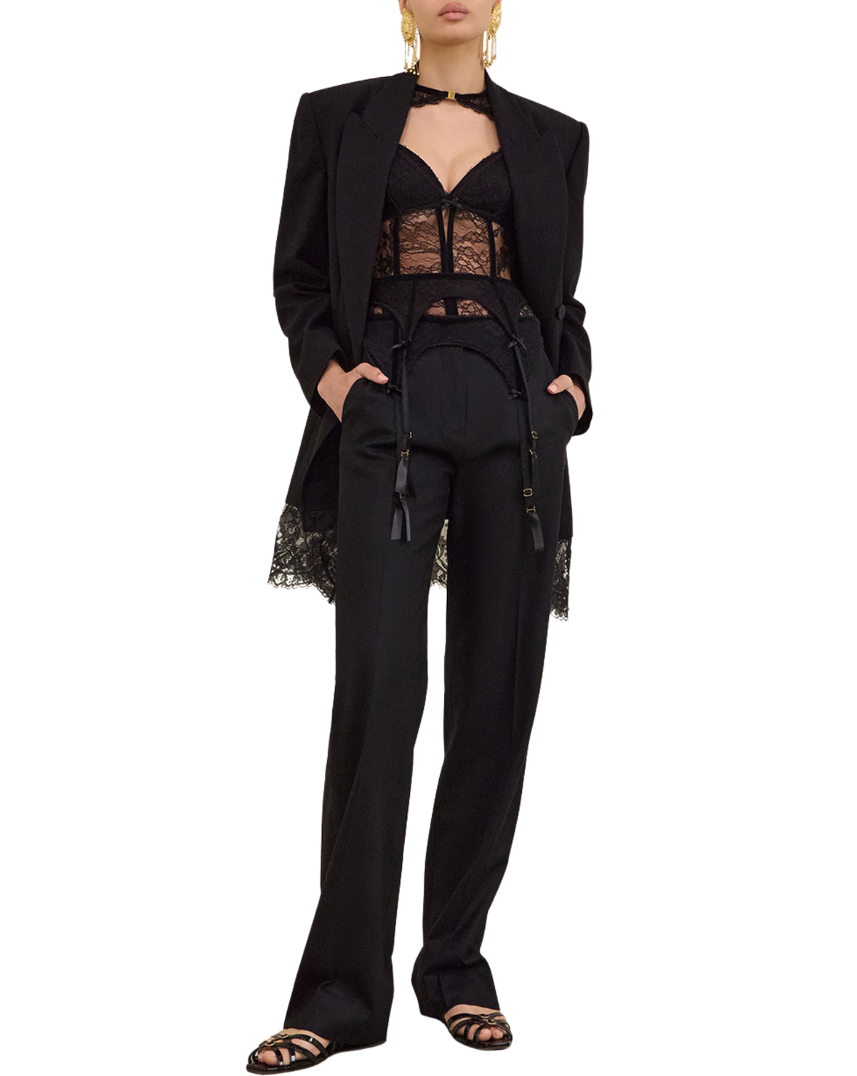 A model wearing a black tailored suit with a lace-trimmed blazer and matching trousers, styled with a black lace bustier and black strappy sandals against a plain white background.