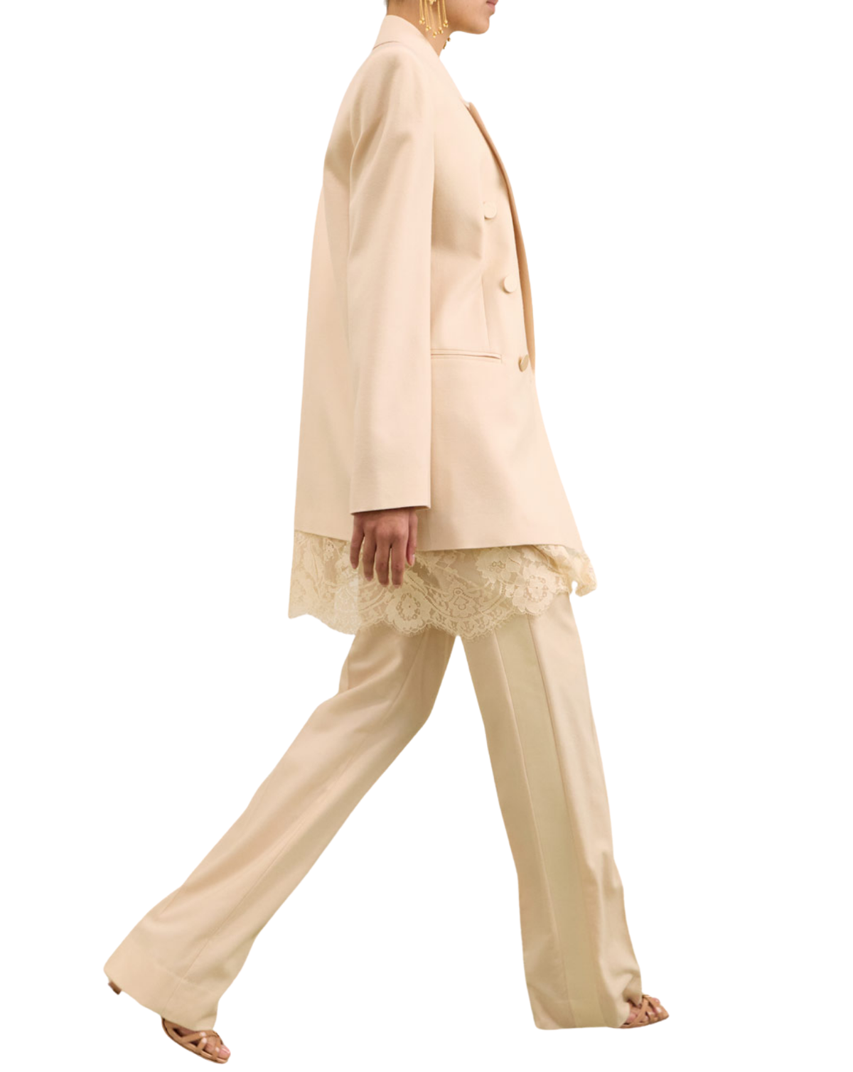A model is shown from the side wearing a light beige double-breasted blazer, matching wide-leg trousers, and a cream lace top, set against a plain white background.
