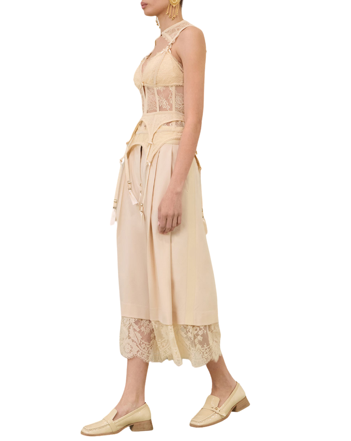 A model is shown from the side wearing a light beige halter-neck midi dress with lace panels, a lace hem, and a matching fabric belt. She is also wearing gold earrings and beige loafers, set against a plain white background.