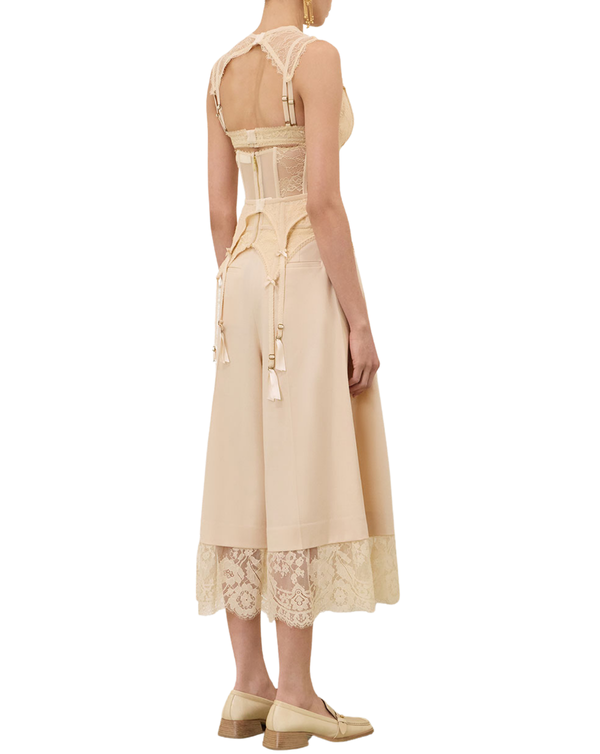 A model shown from the back wearing a beige sleeveless midi dress with lace detailing, a structured corset-style waist, and tassel ties, paired with beige loafers on a white background.