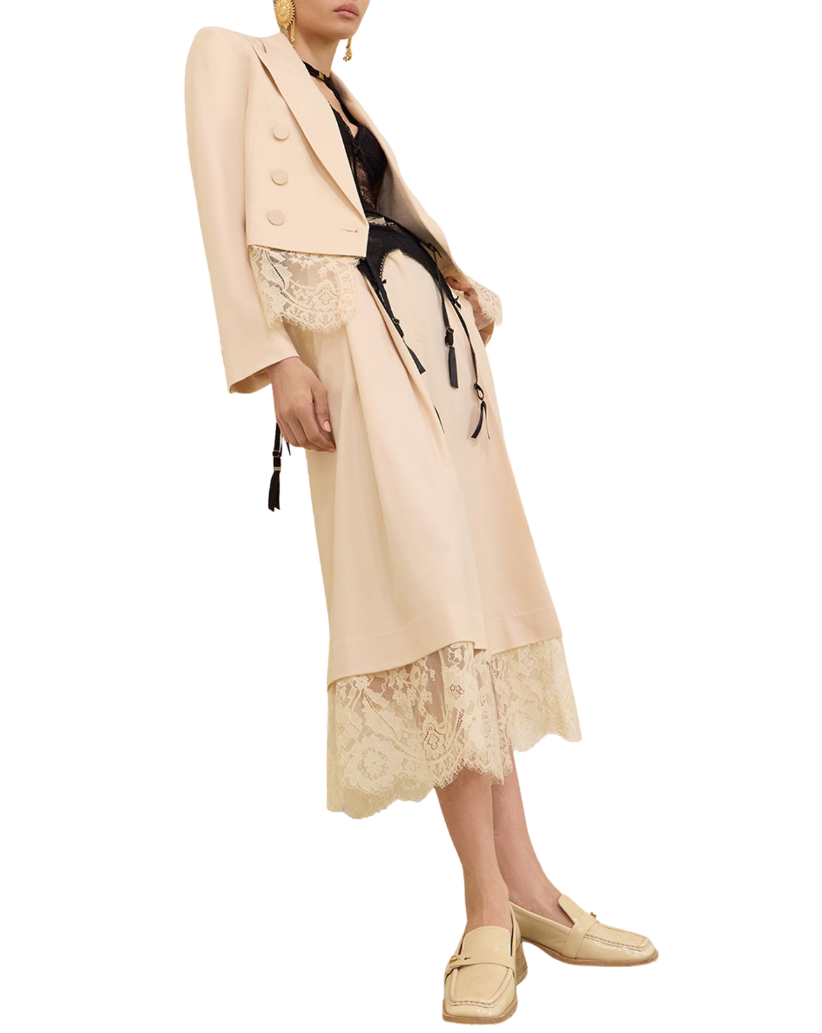 A model is shown wearing a beige two-piece suit consisting of a cropped double-breasted blazer and a matching midi skirt with a lace hem, styled with a black drawstring belt, a black lace top, and beige loafers against a white background.
